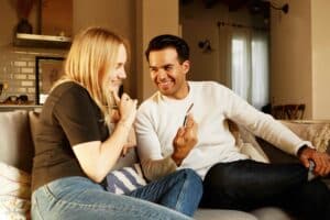 Ein junges Paar sitz auf einer Couch. Spricht glücklich miteinander. Er zeigt ihr etwas auf dem Smartphone. Die Kraft der ehrlichkeit in der Kommunikation -Beziehungstipps . Kommunikation in der Partnerschaft